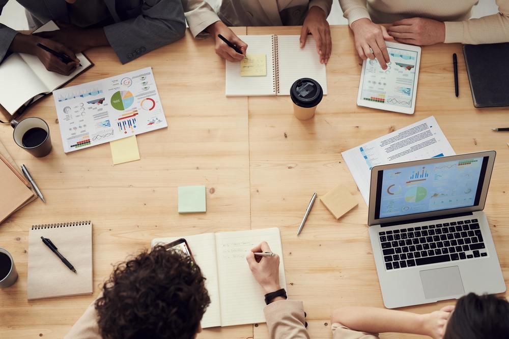 Several people sitting around a table with laptops, ipads , notepads and pens