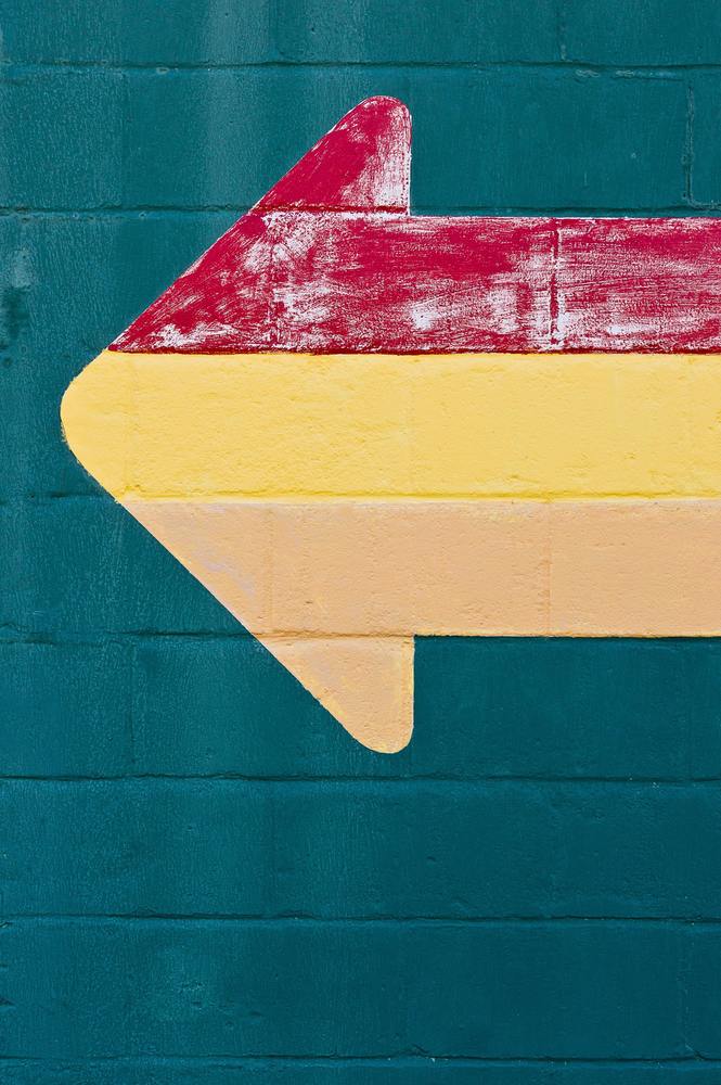 An arrow painted onto a brick wall, the brick wall is green and the arrow is tan, yellow, and red.