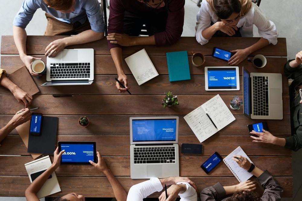 Several people sitting around a table with laptops, ipads , notepads and pens