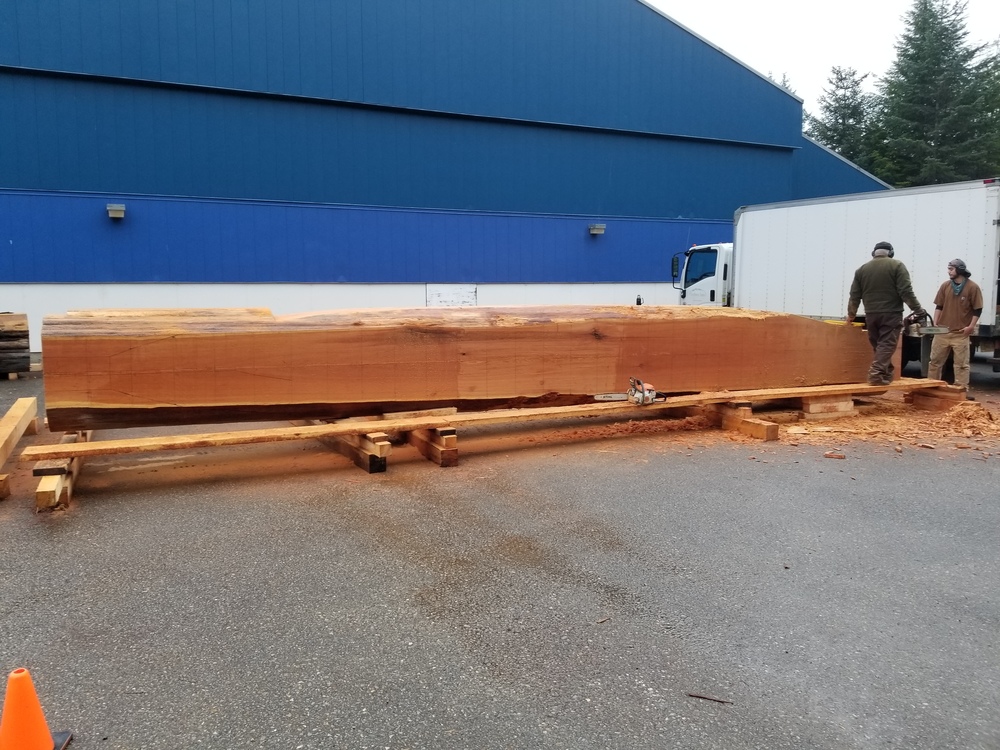Large log in front of the high school building with carving beginning on it and two men near it