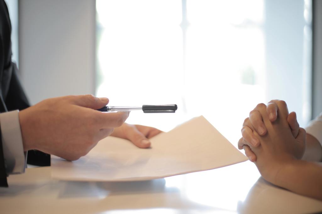 A hand holding a pen and another persons folded hands on a table.